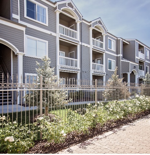 Canyon View Apartments Orem features a modern, multi-story building with gray siding, white trim, balconies, and arched entryways. A metal fence and landscaped garden with shrubs and flowers grace the foreground under a clear sky.