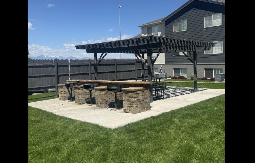 A wooden pergola shades a long picnic table with stone stools on a paved patio in the grassy backyard of Courtside Apartments Orem, bordered by a black fence and a large dark house under a blue sky.