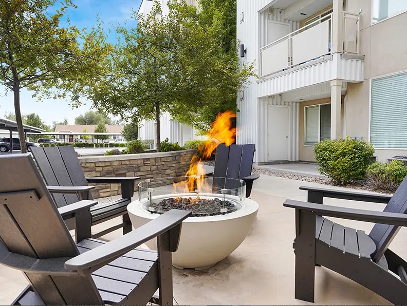 Four dark Adirondack chairs surround a modern round fire pit with a glass barrier at Canyon View Crossing Orem. Flames rise from the center, with a light-colored apartment building and green trees in the background.
