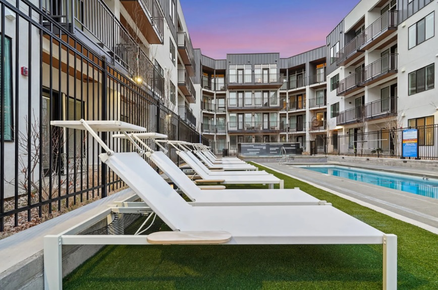 Row of modern white lounge chairs beside a narrow outdoor swimming pool at The Devon at University Place Orem, surrounded by a multi-story apartment building. Purple and pink sunset sky, enclosed by black metal fencing.