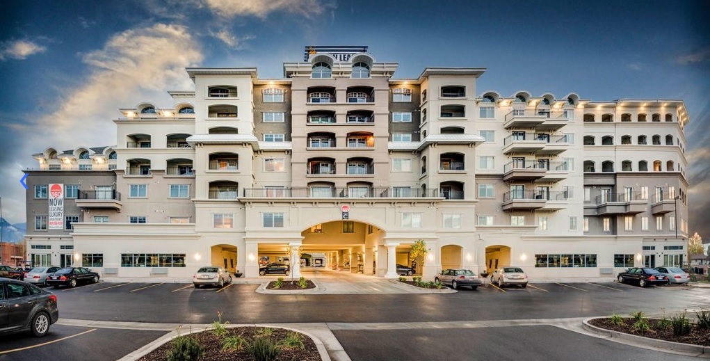 A large, multi-story apartment building with balconies and arched windows, Midtown 360 Orem is lit by soft exterior lighting. Several cars are parked in front on a paved lot under a cloudy evening sky.
