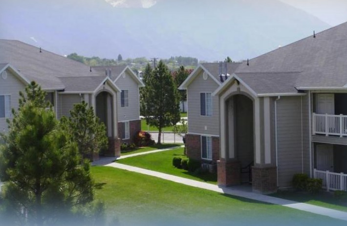 Two beige apartment buildings with balconies face each other at Oakhurst Apartments Orem, separated by green lawns, trees, and sidewalks. Mountains and a hazy sky are visible in the background.