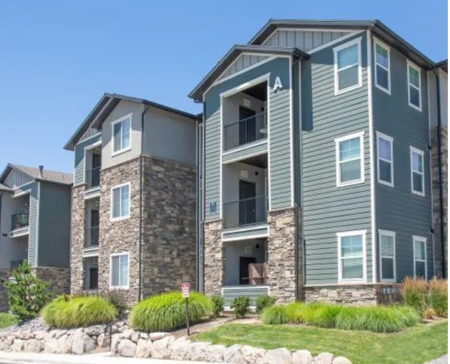 Modern three-story Parc on Center Apartments & Townhomes Orem building with gray siding and stone accents, large windows, balconies, and neatly landscaped grounds with green bushes and grass under a clear blue sky.