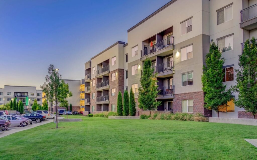 Parkway Lofts Orem is a modern apartment complex with four-story buildings, balconies, and large windows, surrounded by green lawns, trees, and parked cars on a clear evening.