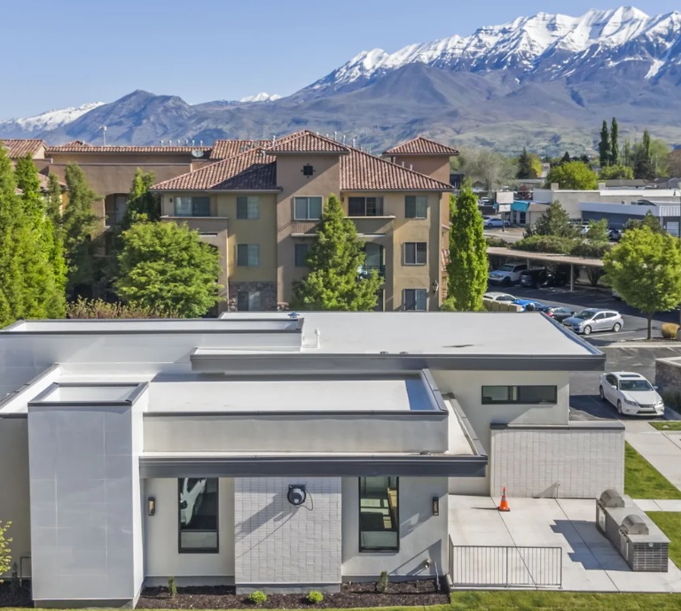 A modern white building with large windows sits in front of the Siena Villas Apartments Orem, surrounded by green trees. Snow-capped mountains and a blue sky create a stunning backdrop.