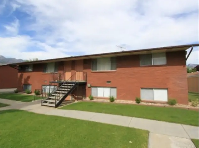 A two-story red brick apartment building at Summertree Orem features a central metal staircase leading upstairs, surrounded by a green lawn and concrete walkway, all beneath a partly cloudy sky.