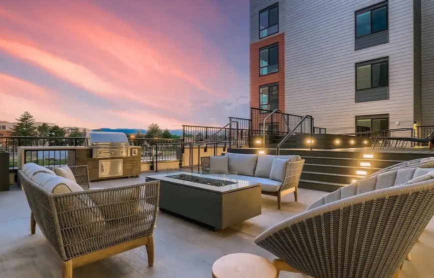 Modern outdoor patio at 281 Willow Fort Collins with cushioned wicker seating, a fire pit table, and a grill, set against a multi-story building at sunset with a colorful sky and stairs featuring built-in lights.