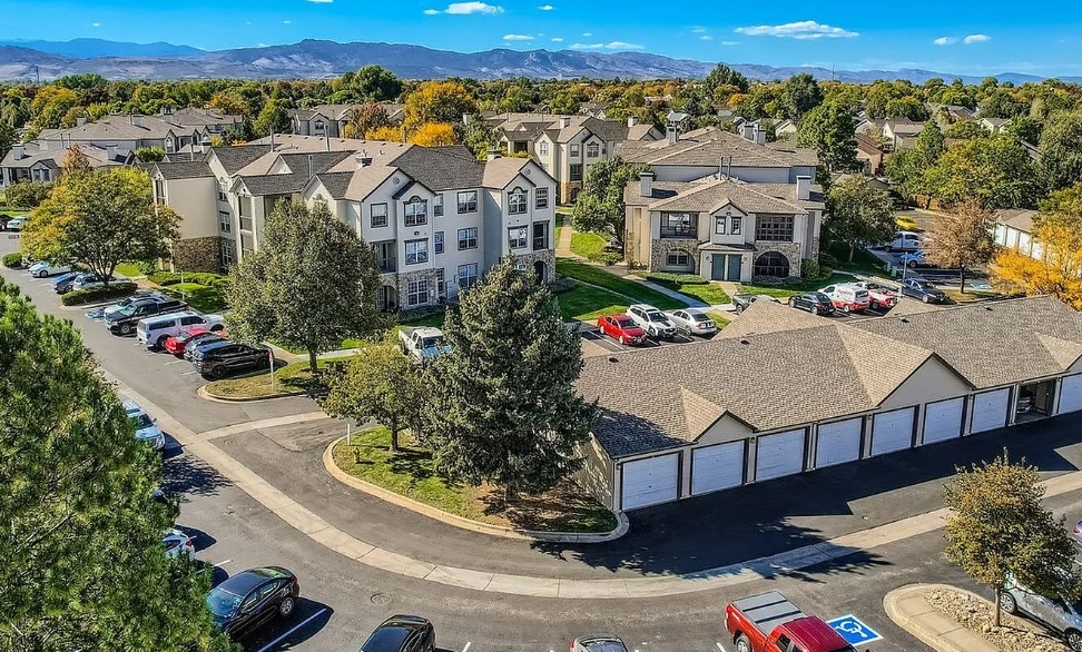Aerial view of Harmony at Willow Springs Fort Collins, a suburban apartment complex with multiple beige buildings, parked cars, garages, and trees with green and autumn foliage. Mountains and a bright blue sky are visible in the background.