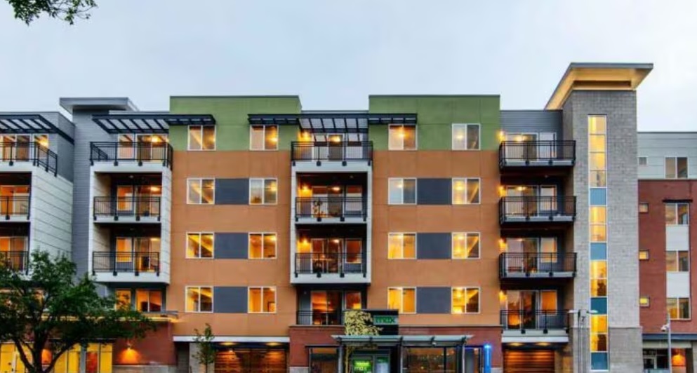 Max Flats Fort Collins is a modern, four-story apartment building with balconies, large windows, and illuminated interiors at dusk. The facade features orange and green panels, with a bus stop and trees in front.