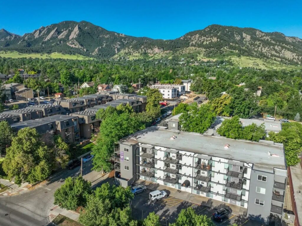 Aerial view of Parker off Pearl Boulder, a mid-rise apartment building surrounded by trees, with more residential buildings nearby and mountains covered in greenery in the background under a clear blue sky.