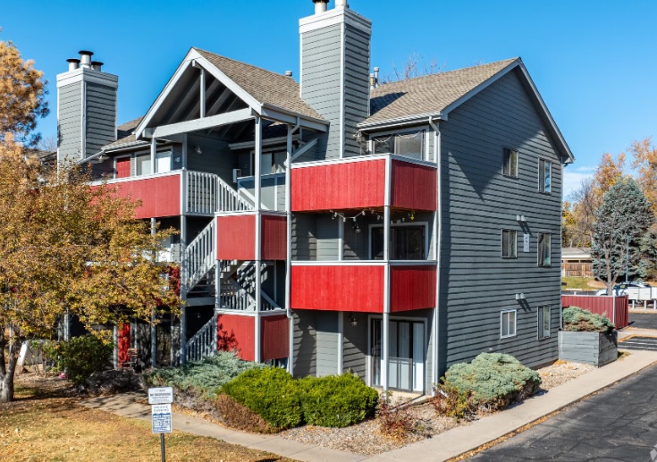 A modern, three-story apartment building at ReNew Foothills Fort Collins features gray siding and red balcony panels, surrounded by shrubs and trees under a clear blue sky.