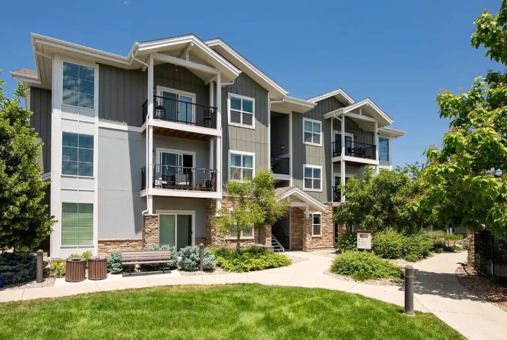 Modern three-story apartment building with gray siding, stone accents, and balconies at The Trails at Timberline Fort Collins. Surrounded by green lawn, trees, bushes, and a curved sidewalk under a clear blue sky.