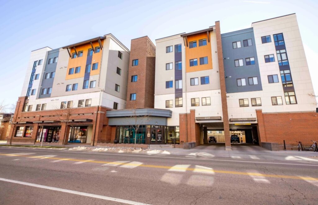 Union on Alley Fort Collins is a modern five-story apartment building with brick, tan, and gray exterior panels, large windows, and ground-floor retail spaces, viewed from across a city street on a sunny day.