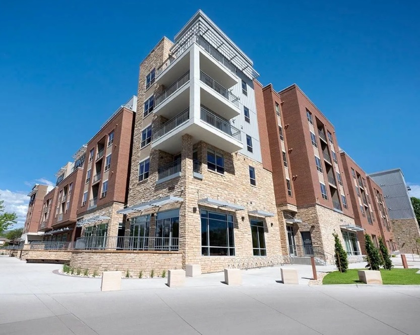 A modern multi-story apartment building, Union on Elizabeth Fort Collins, features a brick and stone exterior, large windows, balconies, and a clear blue sky above. The clean, spacious area offers sidewalks and greenery for a welcoming atmosphere.