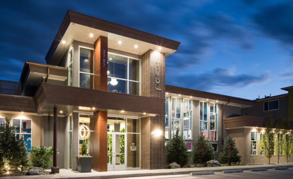 Modern building with large windows illuminated at night, showcasing a well-lit entrance and contemporary architectural design—The Vibe Apartments Fort Collins is surrounded by small trees beneath a clear evening sky with clouds.