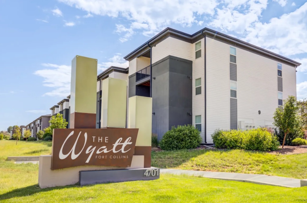 Modern apartment building with white and gray exterior, large windows, and balconies. In front, a contemporary sign reads The Wyatt Apartments Fort Collins with address number 4701. Green grass and shrubs surround the area.