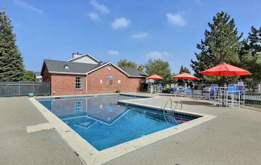 Outdoor swimming pool with clear blue water, surrounded by a concrete deck at Buffalo Run Apartments Fort Collins. Red brick building in the background, patio tables with red umbrellas and chairs on the right, trees and bright sky visible.