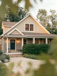 A cozy, beige-colored house on FM Eastwood Court with a covered front porch, white trim, and two upper windows. Bushes and a gravel path lead to the front door, while leafy branches partially frame the image.