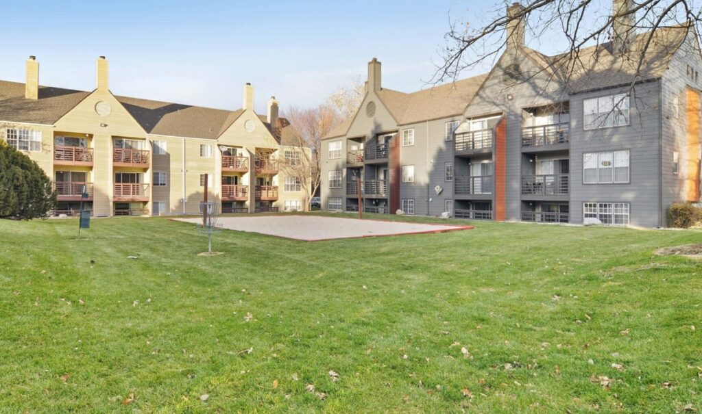 Three-story Habitat at Fort Collins apartment buildings with balconies surround a well-kept lawn and a sand volleyball court under a clear sky. Trees and shrubs are visible around the grassy area in Fort Collins.