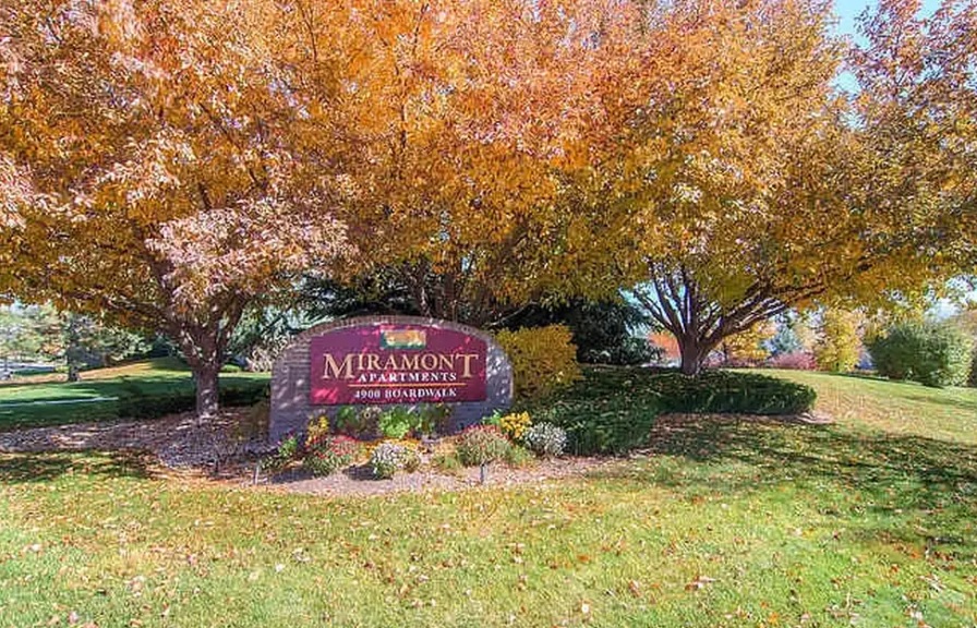 A sign for Miramont Apartments Fort Collins stands surrounded by trees with orange autumn leaves and neatly trimmed grass, set against a clear blue sky in the background.