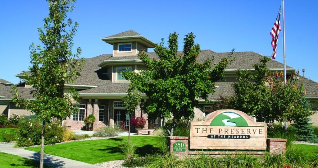 A large, well-maintained building with a sign reading The Preserve at the Meadows Fort Collins stands amid manicured landscaping, trees, and an American flag on a flagpole beneath a clear blue sky.