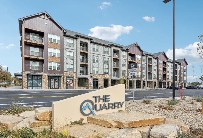 A modern four-story apartment building with balconies and large windows. In the foreground, a stone sign reads “The Quarry Luxury Apartments Fort Collins.” The building faces a paved road under a blue sky with scattered clouds.