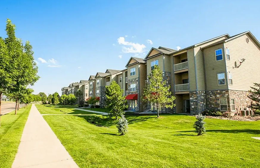 A row of modern, three-story apartment buildings with balconies at Settlers Creek Apartments Fort Collins sits amid well-maintained green lawns and young trees under a clear blue sky, next to a wide sidewalk.