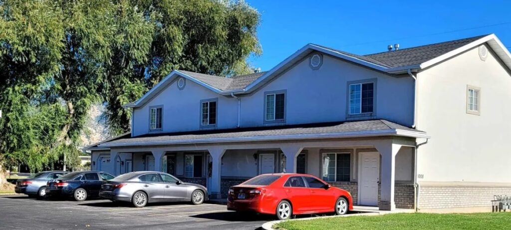 A two-story apartment building at Lanny 1008 Logan with four units, each with its own front door, sits next to a parking lot where several cars—including a red sedan—are parked. Large trees and a clear blue sky complete the scene.