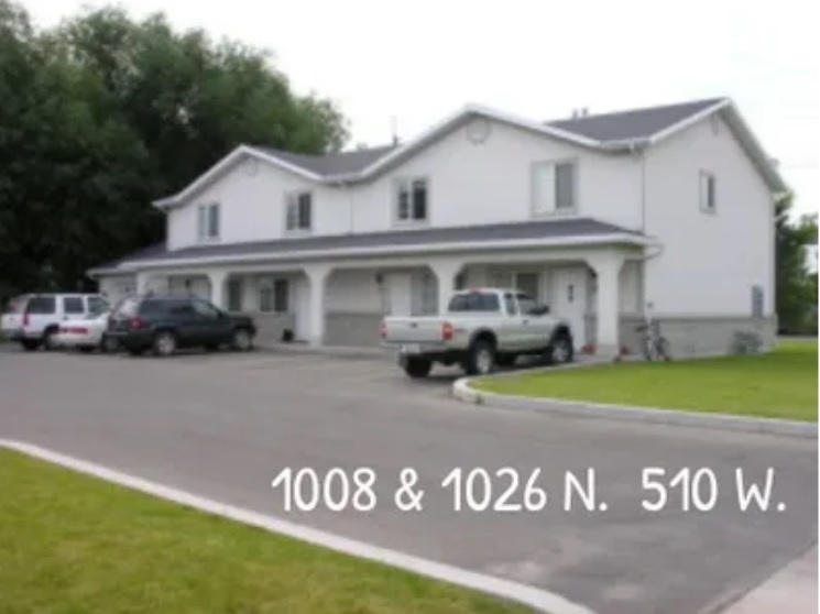 A two-story white duplex with a gray roof stands at 1008 & Lanny 1026 N. 510 W. in Logan. Multiple vehicles are parked in front, and a green lawn with trees surrounds the property. The address appears overlaid in white text.