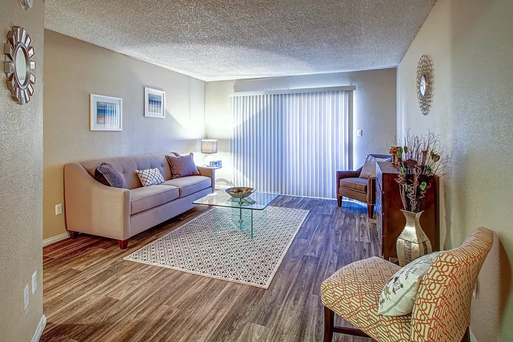 A modern living room at The Marq at 1600 Apartments Las Vegas features a beige sofa, patterned chairs, a glass coffee table on a rug, wooden floors, wall art, and sliding glass doors with vertical blinds welcoming natural light.