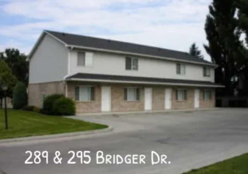 A two-story duplex building with brick and light siding, four white doors, and a large driveway. The address 289 & 295 Bridger Dr. appears at the bottom of the image, referencing Lanny Bridger in Logan. Trees and grass border the building.