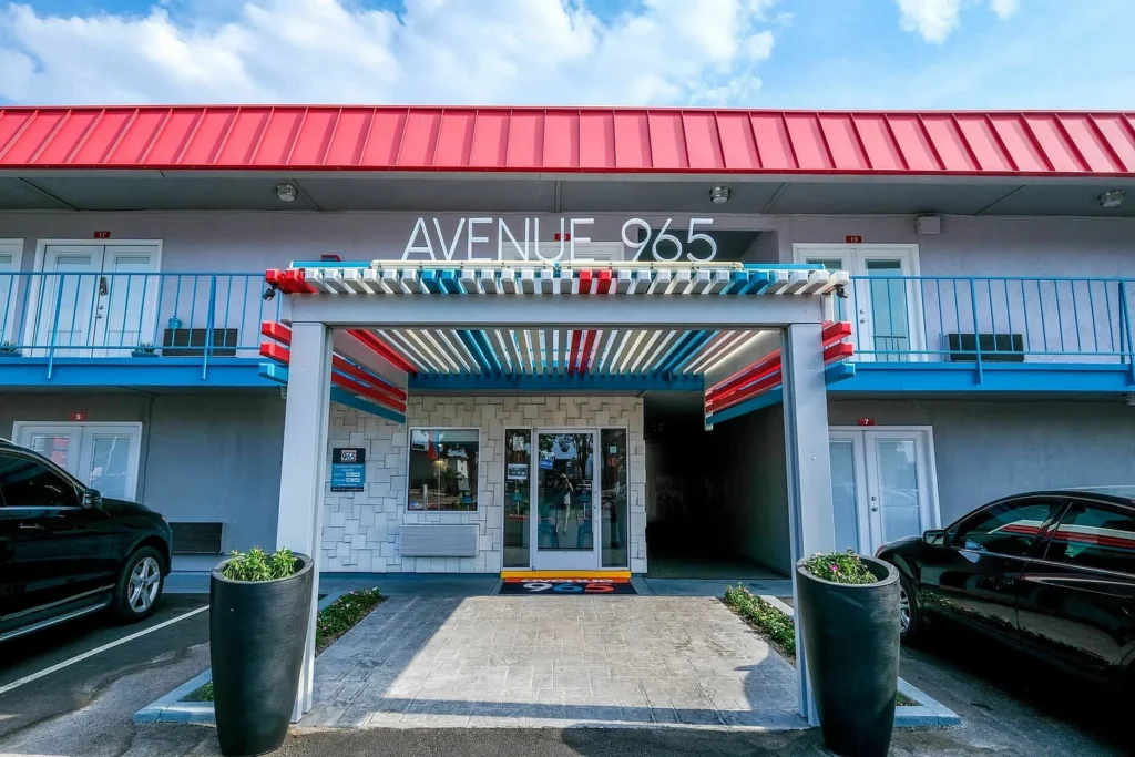 Exterior entrance of Avenue 965 Las Vegas motel with a red, white, and blue canopy, modern design, large planters, and two parked cars on either side of the entrance under a partly cloudy sky.