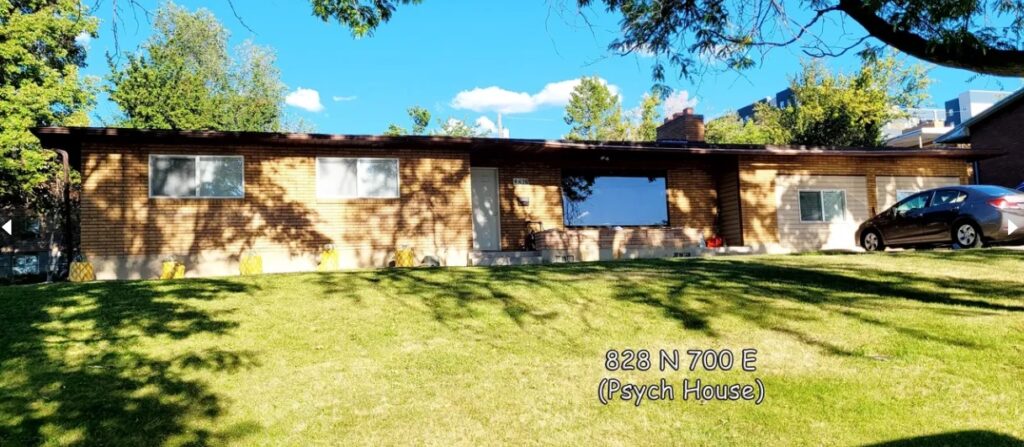 A single-story brick house with large windows and a well-kept lawn, shaded by trees. A gray car is parked in the driveway of this Austin Ridge home at 828 N 700 E (Psych House) in Bennett's Logan neighborhood.