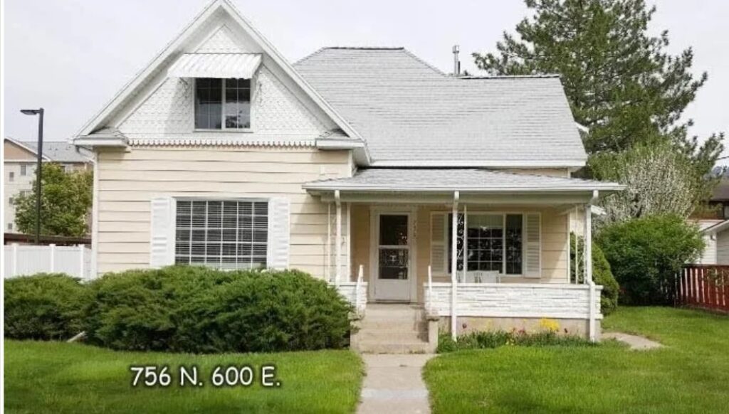 A beige, two-story Austin Ridge Doll House sits behind green bushes and a well-kept lawn in Logan, with white trim and a covered front porch. The address 756 N. 600 E. appears in white text on the lower left.