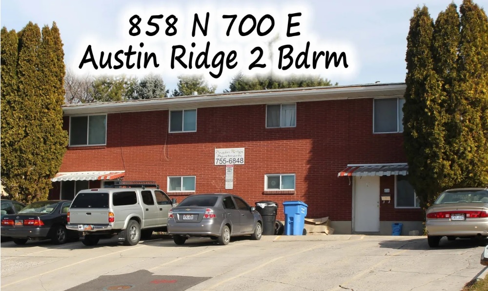 A two-story red brick apartment building with several cars parked in front. Tall evergreen trees line the property. The text reads: 858 N 700 E, Austin Ridge, Logan – 2 Bdrm.