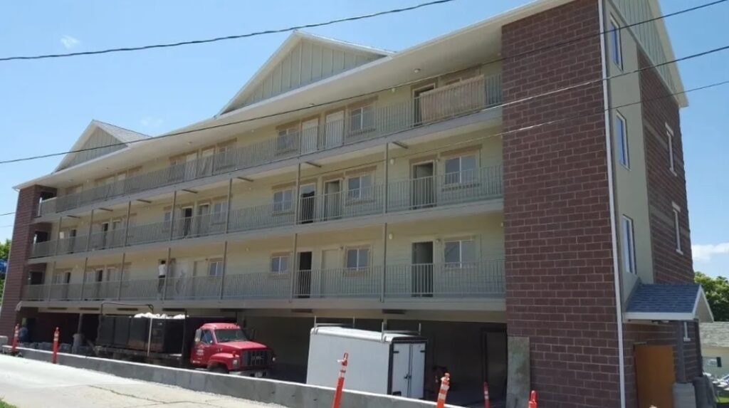 A three-story apartment building at Austin Ridge Meadows Logan features brick and light-colored siding, balconies on each floor, and construction vehicles parked out front. Orange safety cones line the street.
