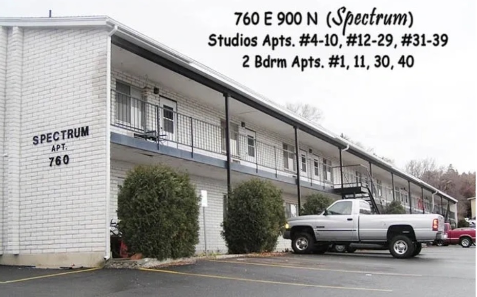A two-story white apartment building with outdoor stairways and railings. A pickup truck is parked in front. The sign lists Spectrum Studio and 1 Bed Logan apartment numbers, along with 2-bedroom units at 760 E 900 N (Spectrum).