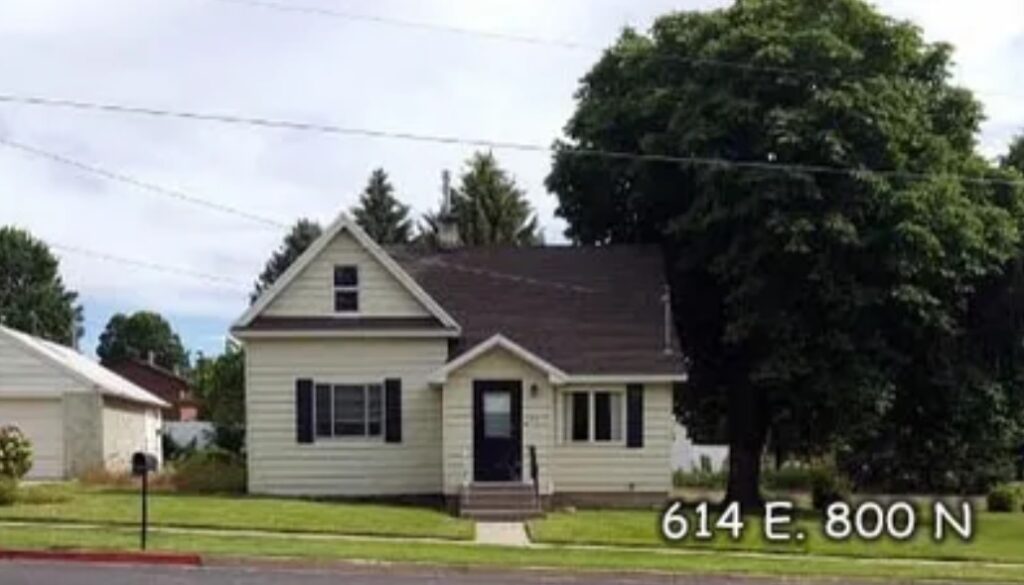 A small, light-colored house with dark shutters and a front porch sits behind a grassy lawn at Austin Ridge. Large trees frame the house, and the address 614 E. 800 N appears in white text at the bottom right.