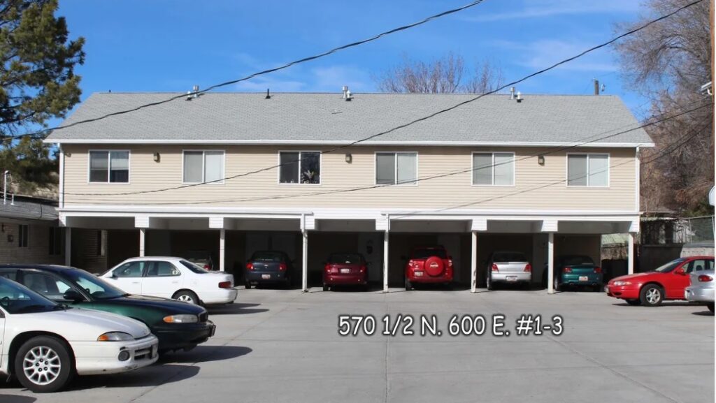 Two-story beige Autumn Hills Triplex Logan apartment building with six covered parking spaces underneath, several cars parked in front, and a blue sky above. The text 570 1/2 N. 600 E. #1-3 is overlaid on the image.