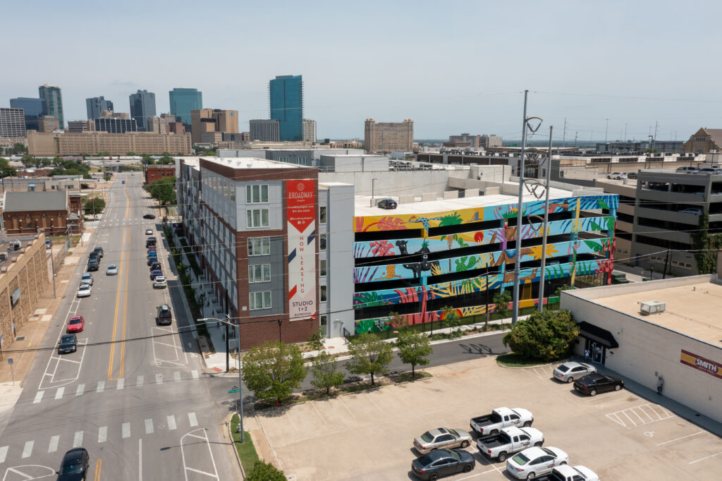 A colorful mural with plants and birds decorates a parking garage wall in an urban area near Broadway Chapter Fort Worth, with city buildings and skyscrapers visible in the background under a clear sky.
