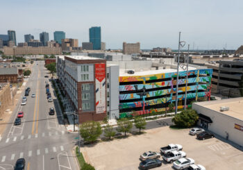 A colorful mural with plants and birds decorates a parking garage wall in an urban area near Broadway Chapter Fort Worth, with city buildings and skyscrapers visible in the background under a clear sky.