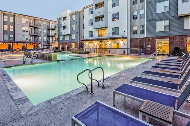 The Arlie Arlington is a modern apartment complex with a large outdoor swimming pool, lounge chairs, and a hot tub area. The buildings feature multiple floors, large windows, and balconies, with the pool area beautifully lit in the evening.