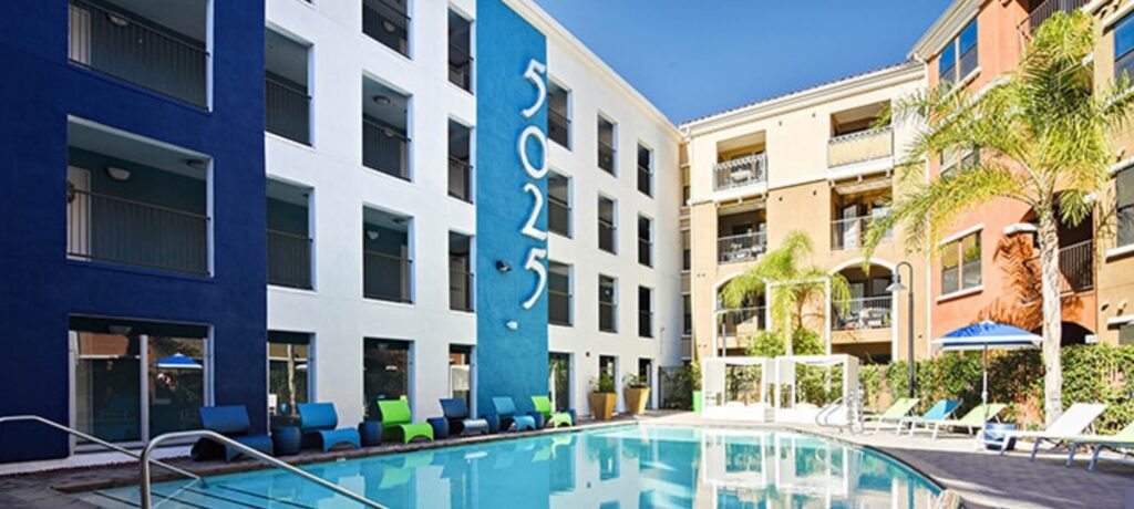 Modern apartment complex with blue and beige exterior walls, large Fifty Twenty-Five numbers on the building, and a swimming pool surrounded by colorful lounge chairs and palm trees on a sunny day.