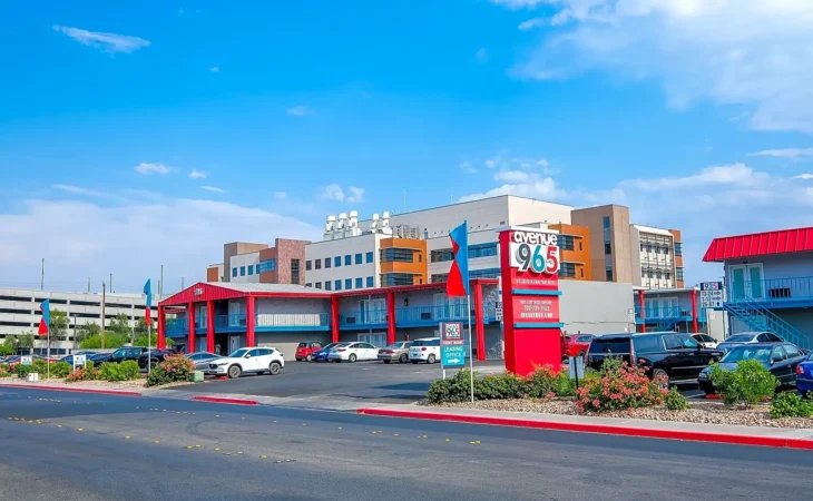 A mid-century style motel with red accents and a sign reading “OYO Hotel 965,” surrounded by parked cars, landscaped bushes, and a larger building and blue sky in the background.