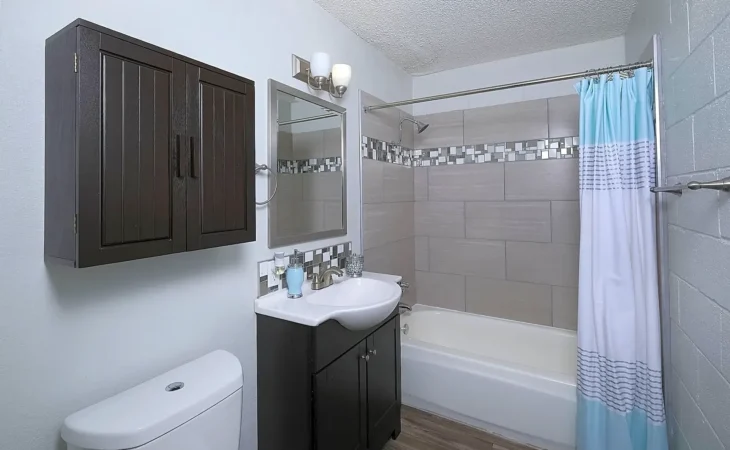 Modern bathroom with a dark wood vanity and cabinet, white sink and toilet, large rectangular wall tiles, a bathtub with a blue and white shower curtain, wood-look flooring, and contemporary lighting above the mirror.