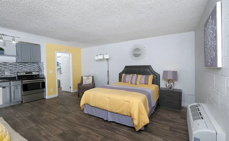A modern studio room with a bed dressed in yellow and gray bedding, a gray armchair, a lamp, wall art, and a kitchen area with gray cabinets and stainless steel appliances on dark wood flooring.