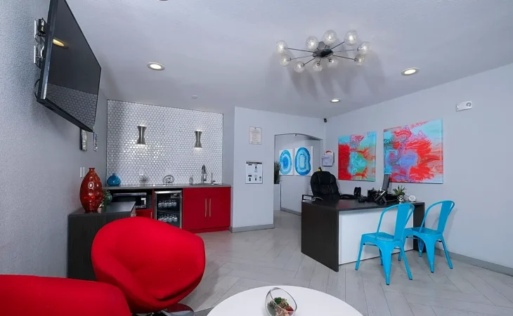 A modern office with gray walls, red chairs, a black desk with two blue chairs, abstract art on the walls, a wall-mounted TV, and a refreshment area with red cabinets and silver decor.