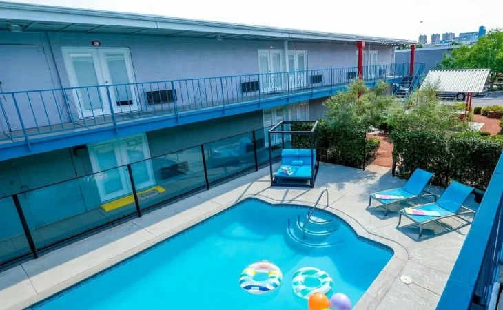 A small outdoor swimming pool with colorful inflatable floats and pool toys, surrounded by a concrete deck with lounge chairs and towels. The pool is enclosed by a fence, with a two-story motel building in the background.