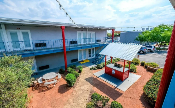 A courtyard with a picnic table, bushes, and a red and blue covered structure is surrounded by a two-story gray building with blue railings and doors. Several cars are parked in the lot outside the courtyard.