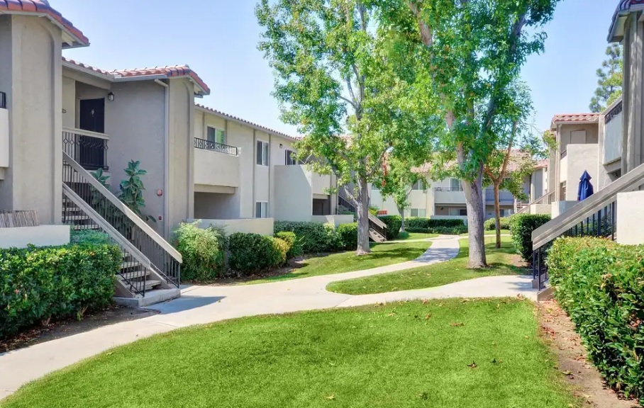 A landscaped apartment complex with light-colored two-story buildings, outdoor staircases, green lawns, bushes, and mature trees, connected by winding concrete walkways under a bright sky.